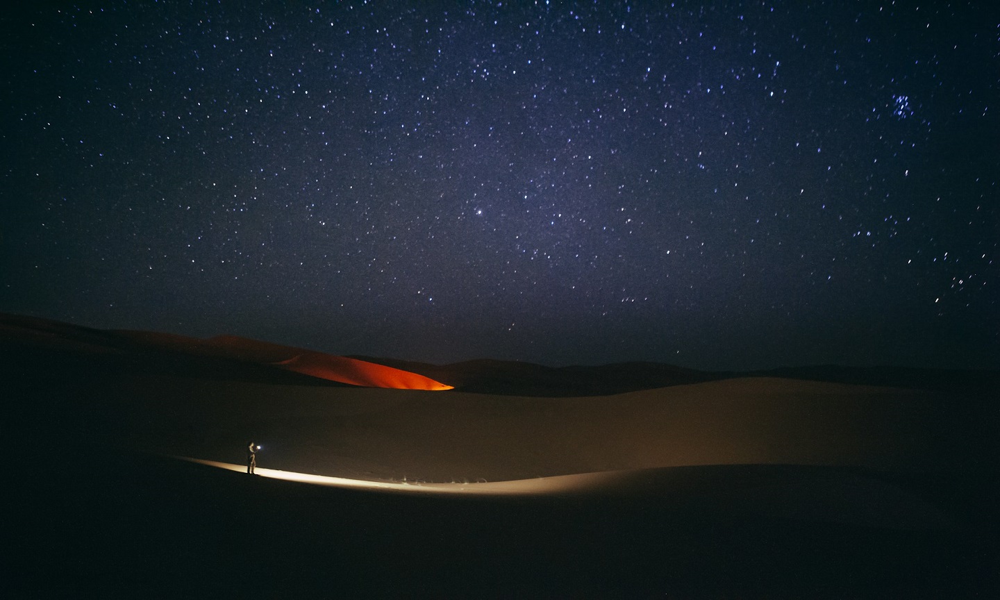A lone person walking across desert dunes beneath a star-filled night sky